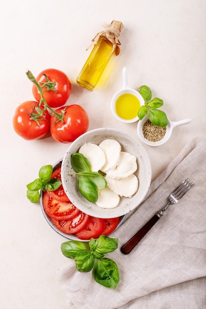 Hausgemachtes Konzept für gesunde Mahlzeiten: Kirschtomaten, Mozzarella-Bällchen, Gewürze, Olivenöl und frisches Basilikum. Mozzarella-Salat über weißer Texturoberfläche. Draufsicht, flach liegen