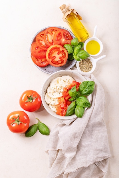 Hausgemachtes Konzept für gesunde Mahlzeiten: Kirschtomaten, Mozzarella-Bällchen, Gewürze, Olivenöl und frisches Basilikum. Mozzarella-Salat über weißer Texturoberfläche. Draufsicht, flach liegen