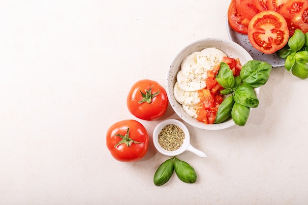 Hausgemachtes Konzept für gesunde Mahlzeiten: Kirschtomaten, Mozzarella-Bällchen, Gewürze, Olivenöl und frisches Basilikum. Mozzarella-Salat über weißer Texturoberfläche. Draufsicht, flach liegen