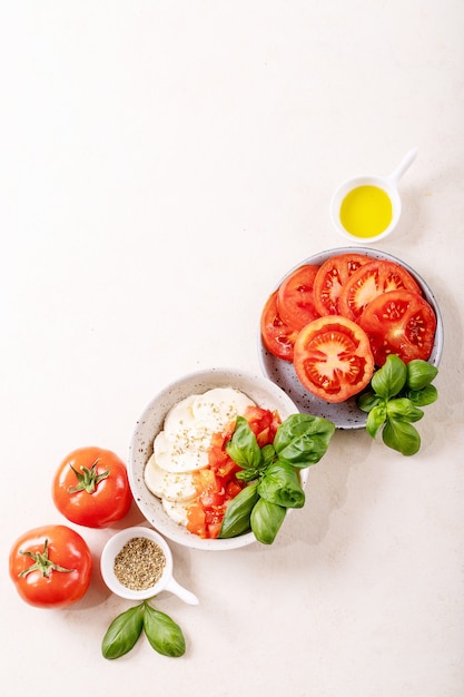 Hausgemachtes Konzept für gesunde Mahlzeiten: Kirschtomaten, Mozzarella-Bällchen, Gewürze, Olivenöl und frisches Basilikum. Mozzarella-Salat über weißer Texturoberfläche. Draufsicht, flach liegen