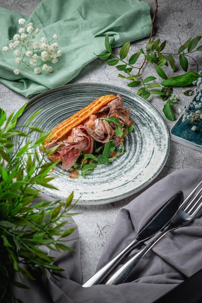 Hausgemachtes Rinderfleisch-Sandwich in Croissant mit Kopfsalat, Tomate und Arugula-Blättern in einem Teller auf einem grauen Steintisch. Speisekarte