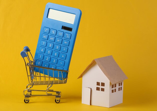 Hauskauf Mini-Papierhaus-Modellrechner im Supermarktwagen auf gelbem Hintergrund