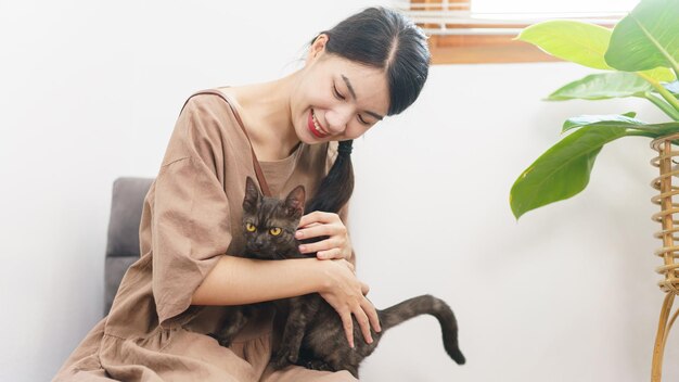 Haustierliebhaber-Konzept Junge asiatische Frau sitzt auf dem Sofa auf dem Boden, um das Fell der Katze zu streicheln und die Katze zu umarmen