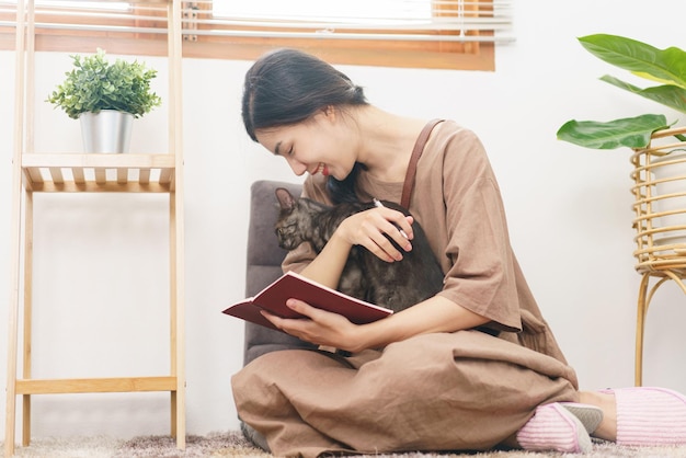 Haustierliebhaber-Konzept Junge asiatische Frau umarmt Katze und macht sich Notizen auf dem Notebook im Wohnzimmer zu Hause