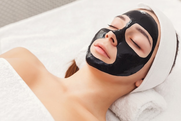 Hautpflege. Porträt des jungen Mädchens mit schwarzer Maske im Gesicht. Reinigende Spa-Behandlung im Schönheitssalon, Kopienraum, Nahaufnahme