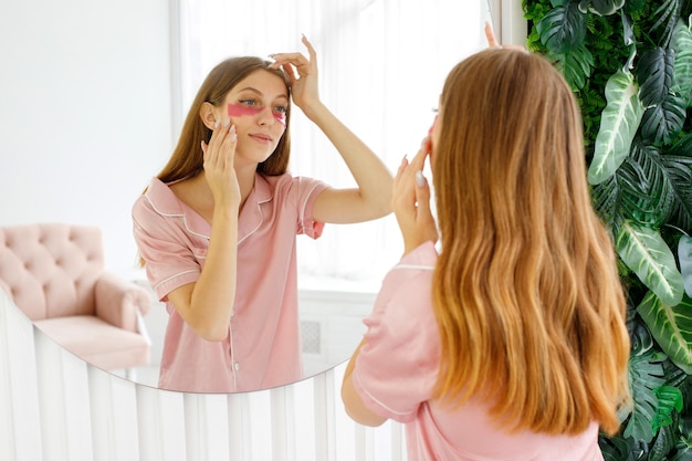 Hautpflegefrau, die Patches unter den Augen anwendet, die sich um die Gesichtshaut kümmern, die zu Hause in der Nähe des Spiegels im Badezimmer steht Junge Frau, die Hautpflege mit modernen Kosmetikprodukten genießt