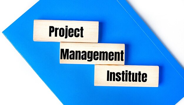 Hay un cuaderno azul sobre un fondo gris claro Arriba hay tres bloques de madera con las palabras Project Management Institute