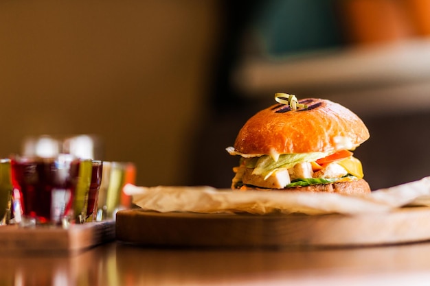 Hay una hamburguesa con pollo, salsa de queso, tomate y verduras en una mesa sobre una plataforma de madera Junto a la hamburguesa hay un conjunto de licores alcohólicos Foto horizontal