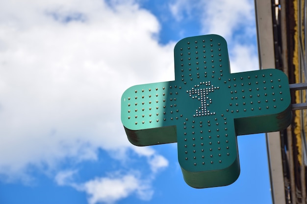 Hay un letrero verde neón en forma de cruz en la pared que anuncia una farmacia u hospital. Contra el fondo del cielo azul con nubes. Hay un lugar a la izquierda para insertar texto.