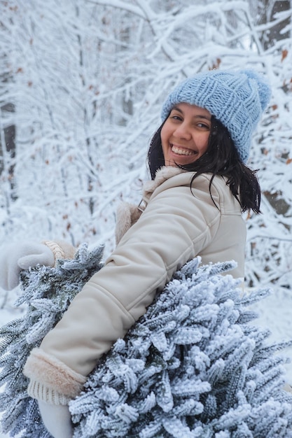 Hay una mujer sonriente sosteniendo un árbol de Navidad cubierto de nieve en el bosque de invierno con una sonrisa en la cara.