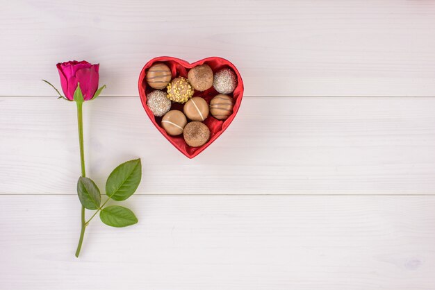 Una hebra de rosa roja y chocolate en una caja en forma de corazón sobre una mesa de madera blanca.