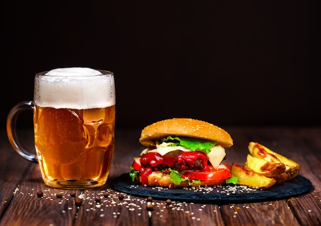 Hecho en casa, deliciosa hamburguesa de ternera con lechuga y patata, un vaso de cerveza servido sobre una tabla para cortar piedra. Oscuro