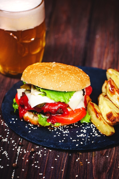 Hecho en casa, deliciosa hamburguesa de ternera con lechuga y patata, un vaso de cerveza servido sobre una tabla para cortar piedra. Oscuro