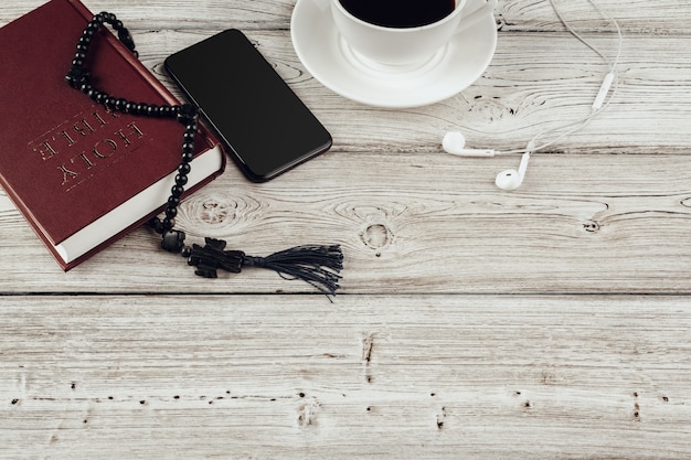 Heilige Bibel und Smartphone mit schwarzer Kaffeetasse auf Holzoberfläche.