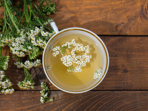 Heilpflanze Schafgarbe, ein Becher mit einer Abkochung von Schafgarbenblüten auf Holzhintergrund