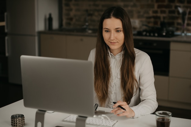 Heimarbeit. Eine kaukasische brünette Frau mit Kopfhörern, die fern an ihrem Laptop arbeiten. Eine Dame in einem weißen Hemd, das Geschäft an ihrem Hauptarbeitsplatz macht.