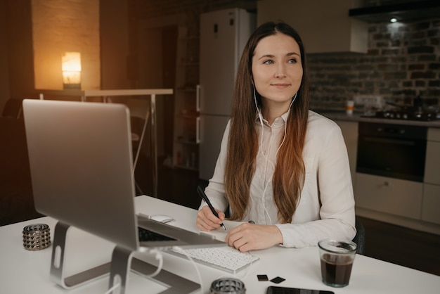 Heimarbeit. Eine kaukasische Frau mit Kopfhörern, die fern an ihrem Laptop arbeiten. Ein glückliches brünettes Mädchen mit einem Lächeln, das Notizen während einer Online-Geschäftsbesprechung an ihrem gemütlichen Heimarbeitsplatz macht.