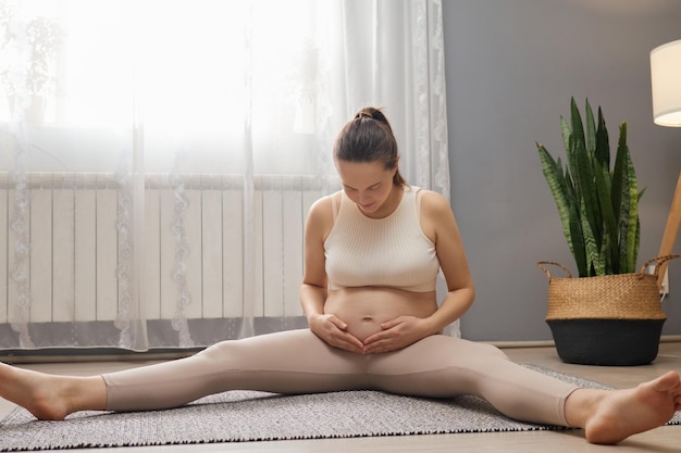 Foto heimtraining während der schwangerschaft vorhersage und bewegung schwangere mit braunen haaren mit pferdeschwanz sitzt auf dem boden und berührt ihren nackten bauch, der sich nach dem training ausruht