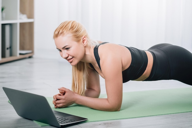 Heimtraining Weibliche Fitness Online-Konferenz Starker Körper Glückliche Frau in schwarzer Sportbekleidung beim Plankentraining grüne Matte mit Laptop im hellen Innenraum