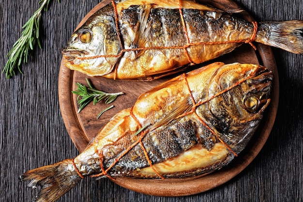 Heiß geräucherte Goldbrasse, Orata-Fisch auf einem runden Holzbrett auf einem dunklen Holztisch mit Gabel, Rosmarin und Pfefferkorn, horizontale Ansicht von oben