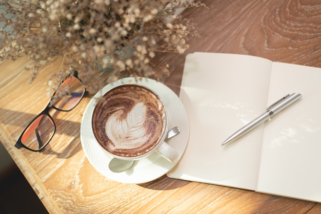 Heiße Kaffee Lattekunst, Anmerkungsbuch und Gläser auf hölzerner Tabelle im Café