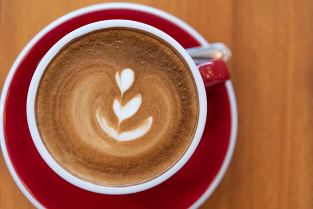 Heiße Kaffeetasse mit Lattekunst in der roten Schale auf hölzernem Hintergrund, Ebenenlage