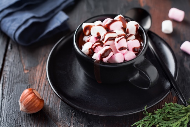 Heiße Schokolade mit Marshmallow-Bonbons in schwarzer Keramikschale auf alter dunkler Holzoberfläche. Selektiver Fokus.