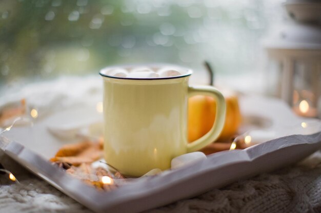 Heiße Schokolade mit Marshmallows in einer gelben Tasse. Weißes Holztablett mit Tasse, Kürbis und Herbstdekorationen in der Nähe des Fensters mit Regentropfen