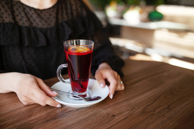 Heiße Tasse Tee mit Frauenhänden im schwarzen Kleid auf Holztisch. Nahaufnahmefoto.