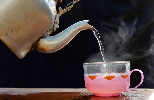 Heiße Teetasse hat heißen Dampf mit Wasserkocher. Auf einem alten Holztisch platziert, schwarzer Hintergrund