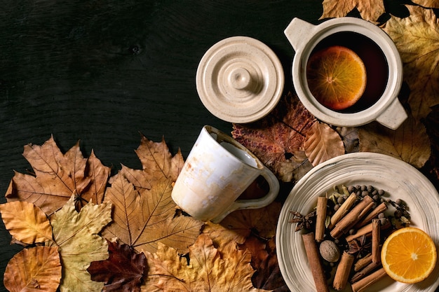 Heißer Glühwein in Keramiktopf und Becher mit Gewürzen, Orange und Herbstlaub auf schwarzem Holzhintergrund. Flach liegen. Gemütliche warme Alkoholgetränke. Flache Lage, Raum