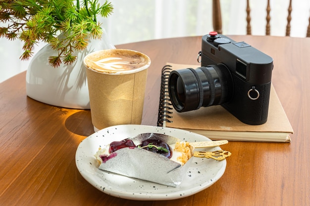 Heißer Kaffee Latte mit Latte Art Milchschaum in einer Tasse aus Papier und Kamera im Notizbuch Blaubeerkuchen auf Holzschreibtisch in der Draufsicht Als Frühstück in einem Café im Café während des Geschäftsarbeitskonzepts