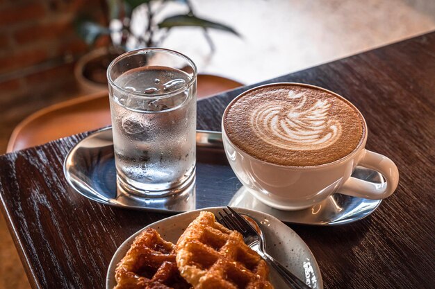 Heißer Kaffee Latte mit Latte-Art-Milchschaum in Schwanenform in Tassenbecher und hausgemachte belgische Waffeln mit Honig auf Holztisch von oben als Frühstück in einem Café im Café während der Geschäftsarbeit