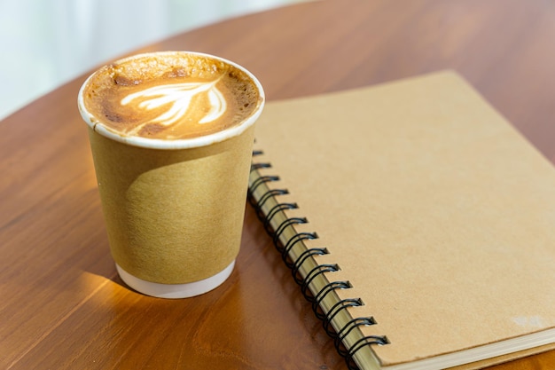 Heißer Kaffee Latte mit Latte Art Milchschaum in Tasse aus Papier und Notizbuch auf Holzschreibtisch in der Draufsicht Als Frühstück in einem Café im Cafeduring Business Work Concept