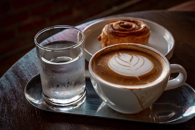 Heißer Kaffee Latte mit Latte-Art-Milchschaum in Tasse und hausgemachtem Rosinen-dänischen weißen Teller auf Holzschreibtisch in der Draufsicht Als Frühstück in einem Café im Café während der Geschäftsarbeit