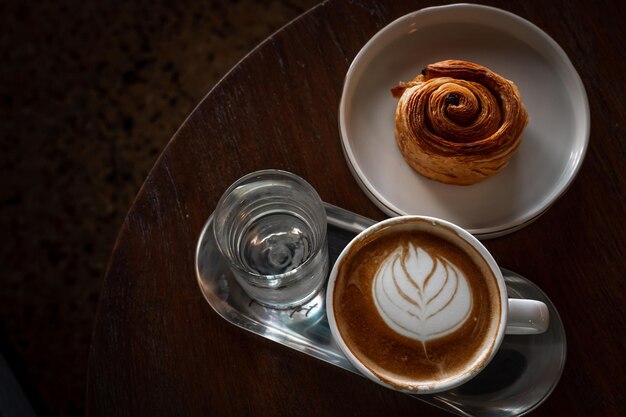 Heißer Kaffee Latte mit Latte-Art-Milchschaum in Tasse und hausgemachtem Rosinen-dänischen weißen Teller auf Holzschreibtisch in der Draufsicht Als Frühstück in einem Café im Café während der Geschäftsarbeit