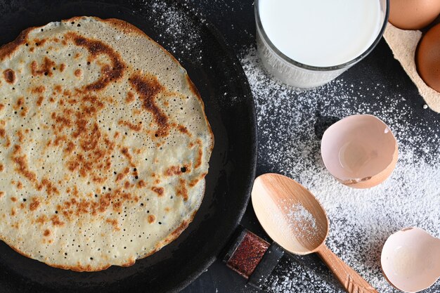 Heißer Pfannkuchen in schwarzer Pfanne auf schwarzem Tisch mit Mehl, Milch und Eiern.