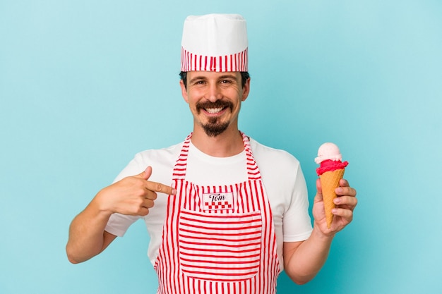 Heladero caucásico joven sosteniendo un helado aislado sobre fondo azul persona apuntando con la mano a un espacio de copia de camisa, orgulloso y seguro