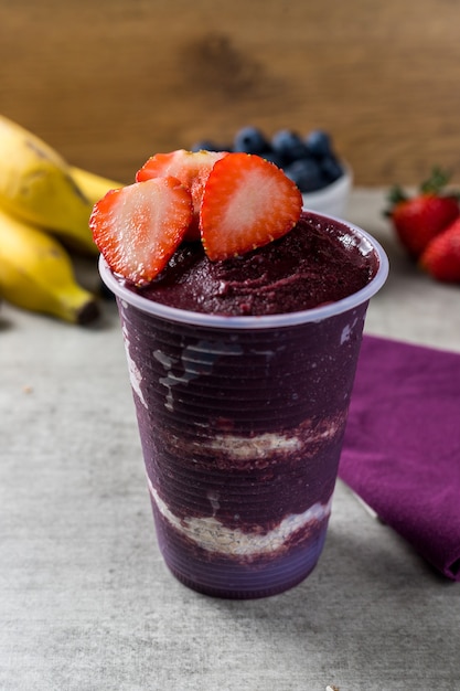 Helado brasileño congelado y de acai berry en un vaso de plástico con hojuelas de fresa y avena. Fondo de plátanos y arándanos. escritorio de madera. Vista frontal del menú de verano.