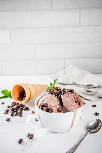Foto helado de café casero, servido con granos de café y hojas de menta, con conos de helado y cucharas en la imagen. fondo de mármol blanco