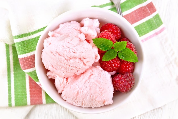 Helado carmesí con bayas de frambuesa y menta en un tazón blanco una cuchara sobre una toalla sobre fondo de tablero de madera desde arriba