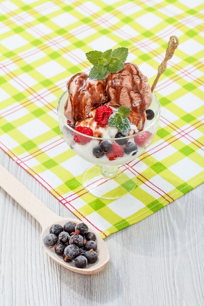 Helado de chocolate y blanco con frambuesa y grosellas y hojas de menta en un vaso sobre una servilleta a cuadros y una cuchara de madera con bayas de grosella congeladas en un escritorio de madera.