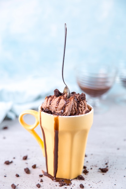 Foto helado de chocolate marrón en una taza y cuchara de color amarillo normal, sobre azul, con cobertura y migas.