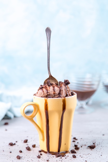 Foto helado de chocolate marrón en una taza y cuchara de color amarillo normal, sobre azul, con cobertura y migas.