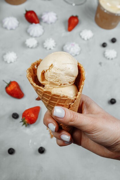 Helado en un cono de waffle sobre un fondo claro en una mano. El helado se está derritiendo. Conos de waffle y fresas