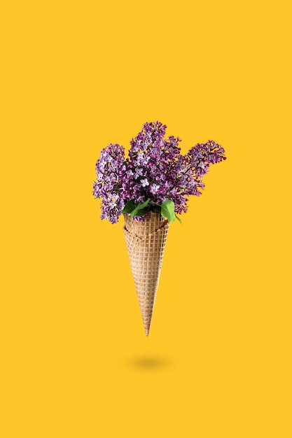 Helado de flores lilas en cono de galleta sobre fondo amarillo desde arriba, hermoso arreglo floral. Flores púrpuras cayendo o volando en movimiento. Día de la madre, 8 de marzo, día de San Valentín, cumpleaños