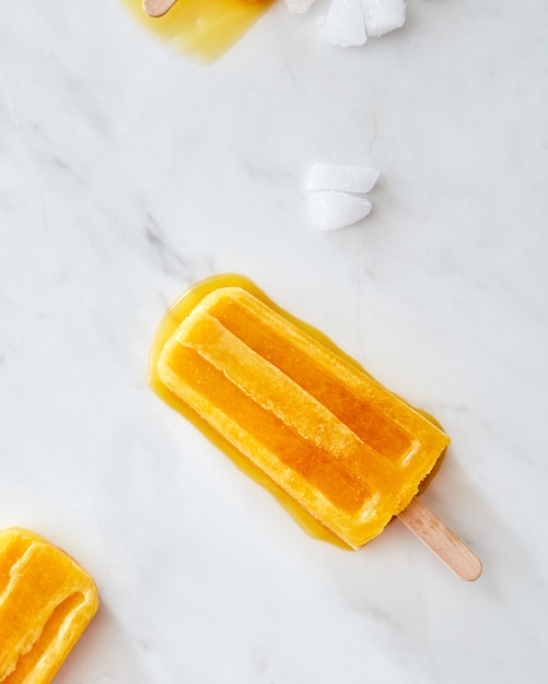 Helado de frutas de naranja en un palito con rodajas de hielo sobre un fondo de mármol gris con espacio para texto. El concepto de postre de verano. Endecha plana