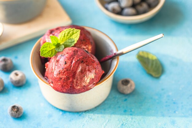 Helado de plátano vegano casero o helado con arándanos en un recipiente sobre fondo azul. Postre saludable.