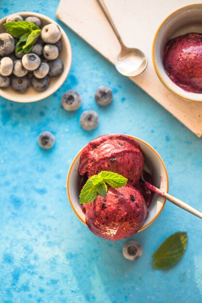 Helado de plátano vegano casero o helado con arándanos en un recipiente sobre fondo azul. Postre saludable.