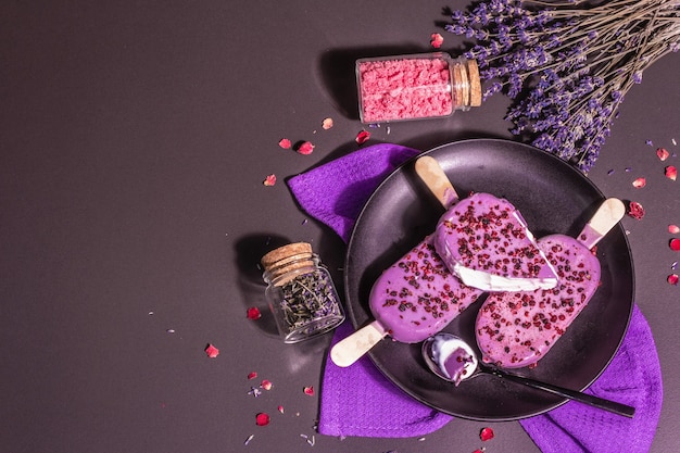 Helado con sabor a lavanda. Ramo de flores, azúcar rosa, pétalos de rosa. Luz dura de moda, sombra oscura. Fondo de hormigón de piedra negra, vista superior